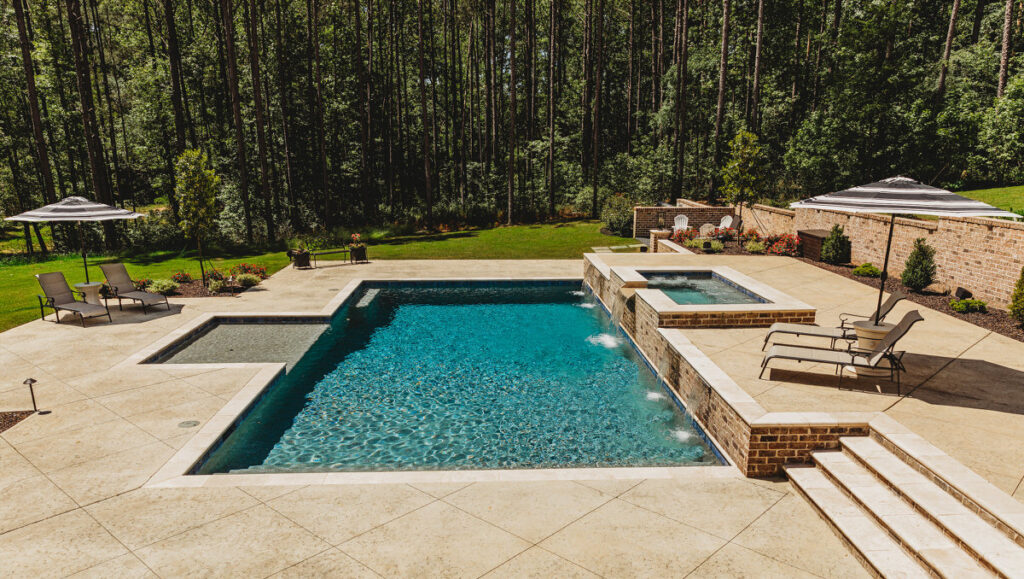 Custom gunite pool with integrated spa, stone coping, concrete deck, lounge chairs, and umbrellas in wooded backyard setting