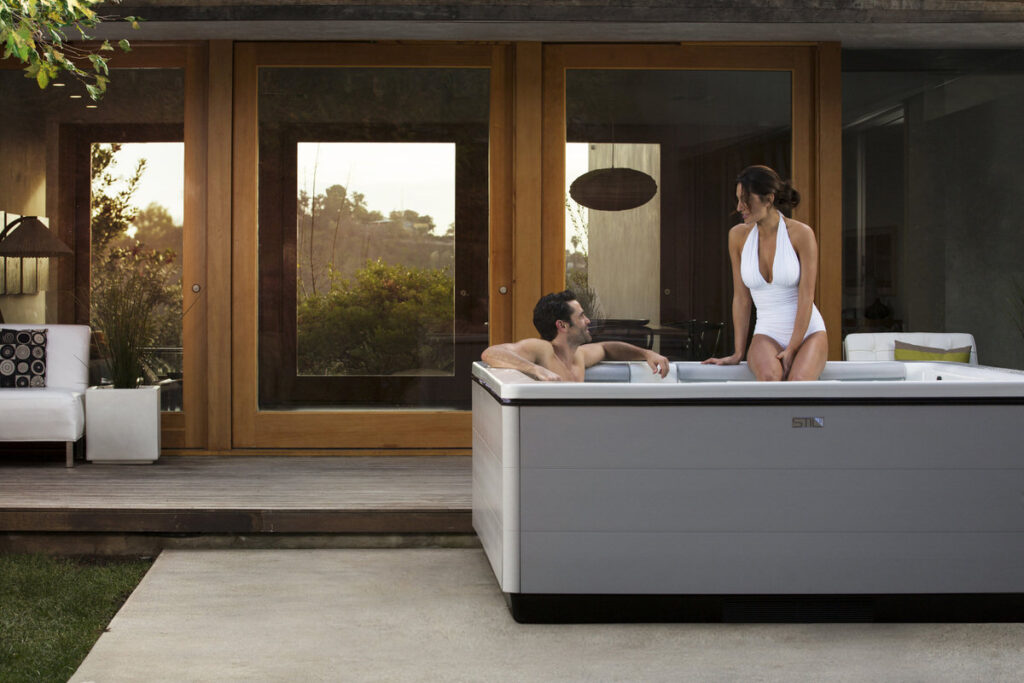 Modern hot tub on wooden deck with couple relaxing, white outdoor furniture visible, contemporary home setting
