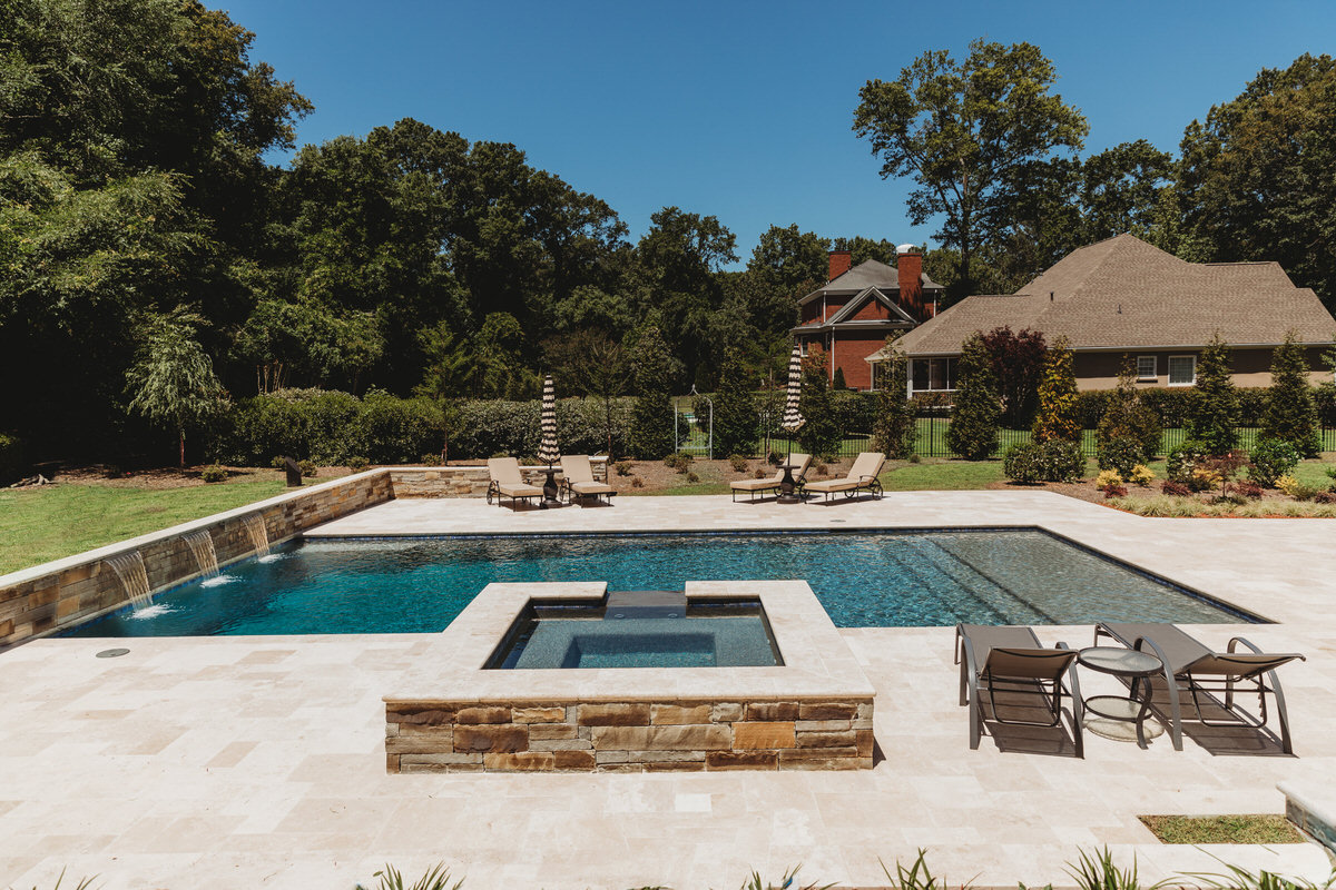 Custom gunite pool with integrated spa, stone coping, concrete deck, lounge chairs, and umbrellas in wooded backyard setting