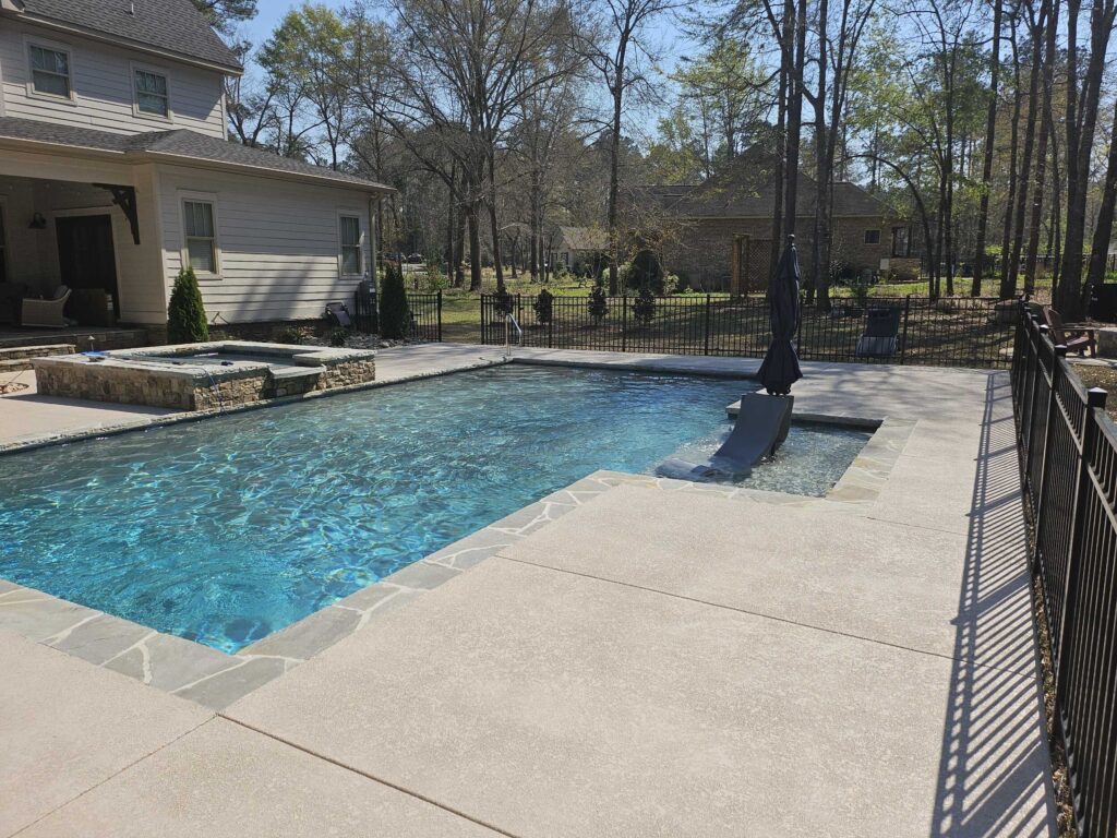 Ground-level view of a crystal clear inground pool with an attached stone hot tub and black iron safety fence surrounding a Georgia backyard
