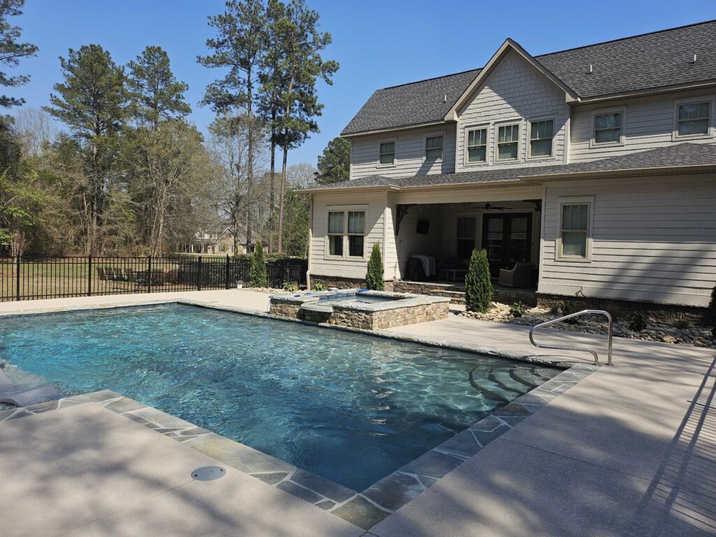 Inground pool with clear blue water and stone-trimmed attached spa next to a covered porch and two-story gray home in Middle Georgia