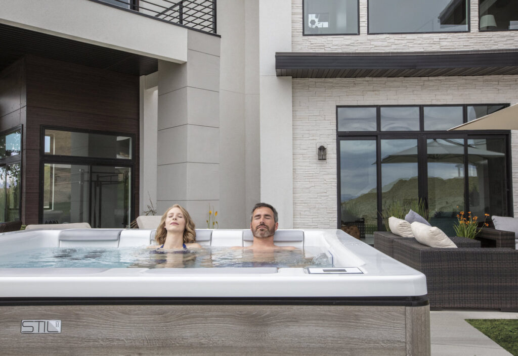 Couple relaxing in square hot tub with modern home architecture and outdoor seating area in background