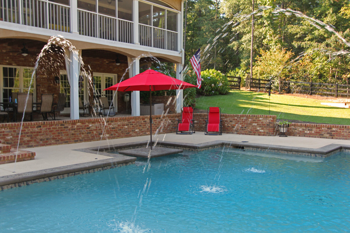 Red Galtech patio umbrella providing shade over poolside lounge chairs with water features and brick retaining wall