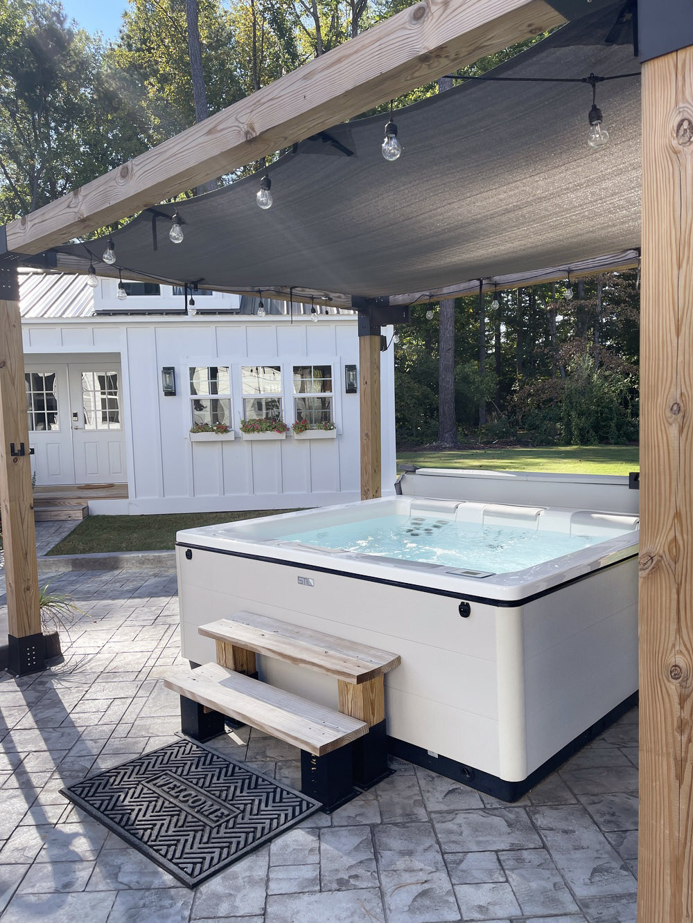 White hot tub installed under wooden pergola with string lights on paver patio next to white shed in backyard